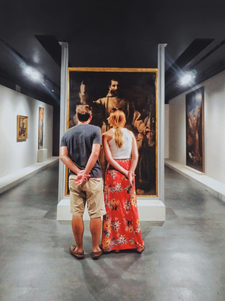 A couple stands inside an art gallery in Brazil, engaging with classical paintings.